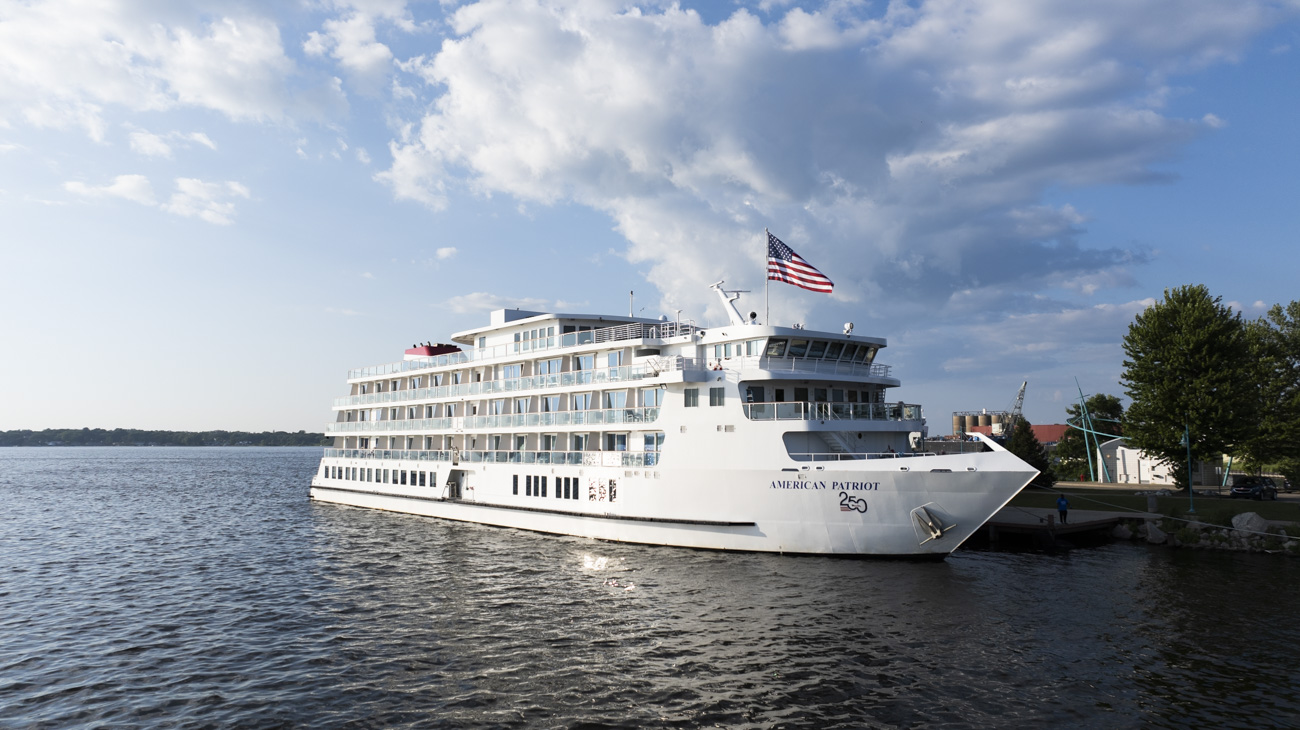 A large white cruise ship with an American flag docked at a pier on a sunny day, with blue skies and scattered clouds in the background.