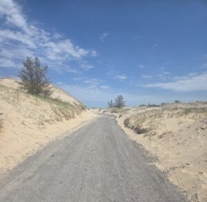 A gravel path runs between sandy dunes with sparse grass and a few trees, beneath a blue sky with scattered clouds.