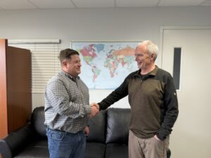 Two men are standing in an office, smiling and shaking hands in front of a black couch. A world map hangs on the wall behind them, and blinds cover a window to the left.