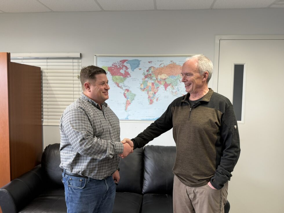 Two men are standing in an office, smiling and shaking hands in front of a black couch. A world map hangs on the wall behind them, and blinds cover a window to the left.