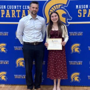 A girl in a red dress holds a certificate and stands next to a man in a white shirt, both smiling, in front of a blue Mason County Central Spartans backdrop.