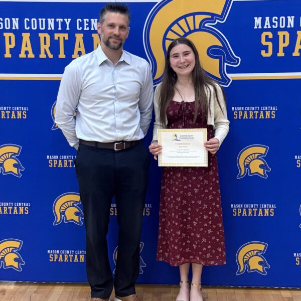 A girl in a red dress holds a certificate and stands next to a man in a white shirt, both smiling, in front of a blue Mason County Central Spartans backdrop.